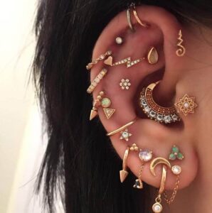 Left ear covered in gold earrings of various shapes and sizes, including hoops, studs, and gemstone drops across multiple piercings.
